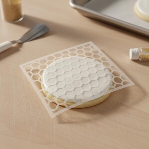 May include: A round sugar cookie with white icing, decorated with a honeycomb pattern using a stencil. A spatula, sprinkles, and a baking sheet are in the background. The cookie is on a light wooden surface.