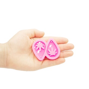 May include: Pink silicone mold for making resin earrings. The mold features two teardrop-shaped cavities with a marijuana leaf design.