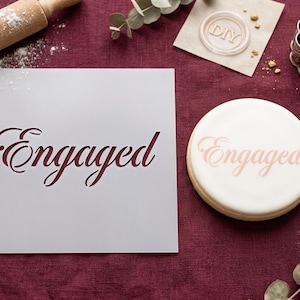 May include: A white stencil with the word "Engaged" in a cursive font is displayed on a burgundy surface. A round cookie with the same word is next to the stencil. A rolling pin, cookie cutters, and a knife are also visible.
