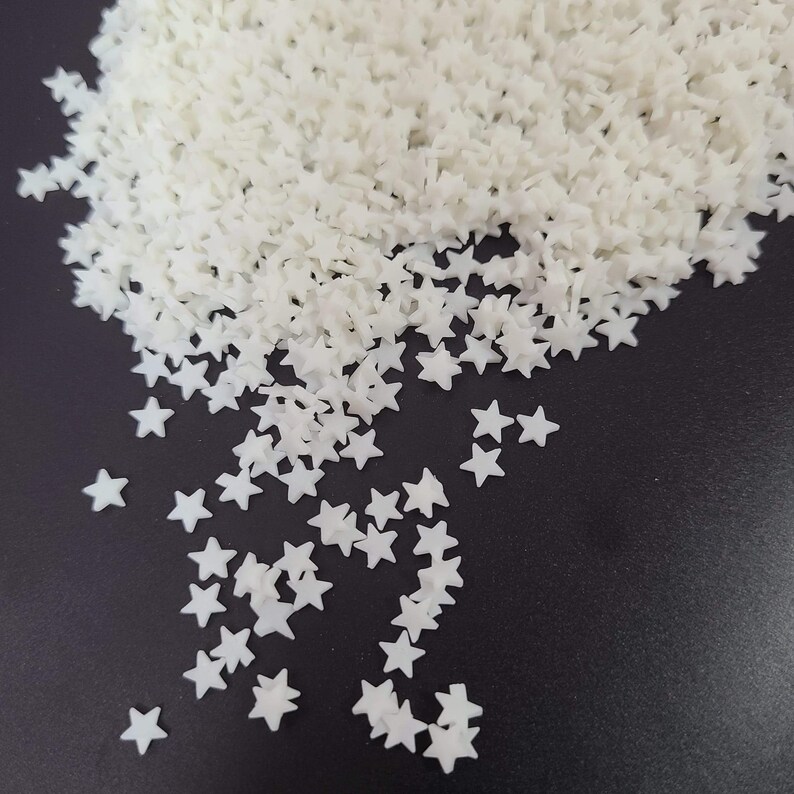 May include: A pile of small, white star-shaped embellishments. The stars are scattered across a dark surface, with a larger concentration of them in the upper portion of the image. The stars appear to be made of a solid material.