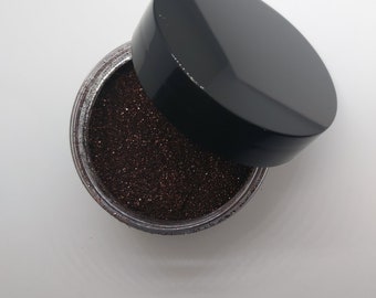 May include: A small jar of brown glitter with a black lid. The lid is off and the glitter is visible.