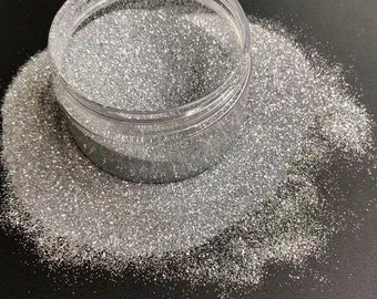 May include: A clear plastic jar filled with silver glitter. The glitter is loose and spills out of the jar onto a black surface.