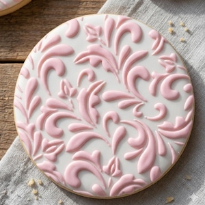 Pode incluir: Um biscoito redondo com uma base branca e um design floral rosa em relevo. O biscoito tem uma borda marrom clara e está colocado sobre uma superfície de madeira com um pano cinza. O design apresenta folhas e espirais estilizadas.