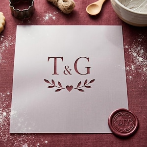 Può includere: Uno stencil quadrato semitrasparente con le lettere T & G in carattere serif, centrate sopra un grafico decorativo di due rami frondosi simmetrici che fiancheggiano un piccolo cuore. Include un sigillo in ceralacca bordeaux con DIY.