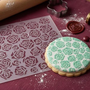 May include: A decorated sugar cookie with a green rose pattern, a clear stencil with a rose design, and baking tools. The cookie is round with a scalloped edge. A rolling pin and a wax seal are also visible.