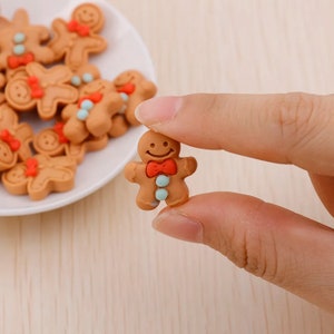 May include: A gingerbread man cookie charm with a red bow tie and blue icing details. The charm is made of brown, red, and blue plastic and is about 1 inch tall.