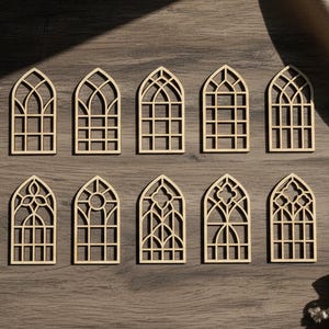May include: Ten laser-cut wooden window ornaments in various Gothic arch designs. The ornaments are light brown and arranged on a dark wood surface. Each window features intricate latticework and decorative details, ideal for crafting or home decor.