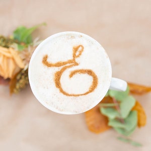 K&ouml;nnte beinhalten: Eine wei&szlig;e Keramiktasse mit einem Zimtmuster in Form des Buchstabens "G" auf dem Schaum.