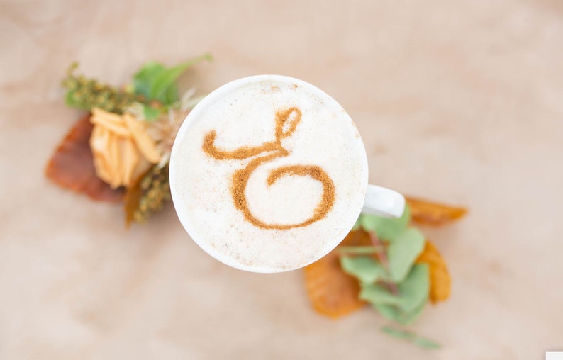 K&ouml;nnte beinhalten: Eine wei&szlig;e Keramiktasse mit einem Zimtmuster in Form des Buchstabens "G" auf dem Schaum.