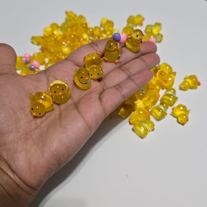 May include: A selection of small, translucent yellow duck figurines, some with painted details. Some ducks are holding balloons. The figurines are scattered on a white surface, with some held in a hand.