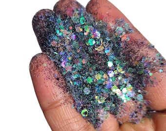May include: A close-up of iridescent glitter in various sizes and shapes, including hexagons, circles, and stars. The glitter is a mix of black, green, blue, and purple colors.