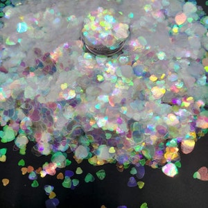 Può includere: Primo piano di glitter a forma di cuore iridescente in una varietà di colori, tra cui bianco, rosa, verde e blu. Il glitter è sparso su una superficie nera.