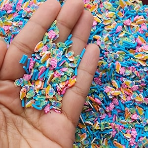 May include: A close-up of a hand holding a variety of colourful fish-shaped and stick-shaped sprinkles. The sprinkles are made of polymer clay and are perfect for decorating cakes, cupcakes, and other desserts.