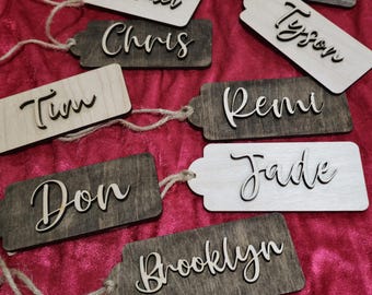 May include: A set of ten wooden gift tags with names written on them in a cursive font. The tags are various shades of brown and white and have twine attached to them.