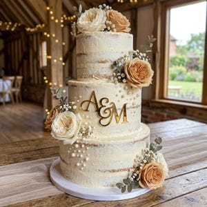 Può includere: Una torta a tre piani semi-nuda con glassa bianca, decorata con rose color crema e marrone chiaro, piccoli fiori bianchi, foglie di eucalipto e perle bianche. Un topper per torta in acrilico dorato con le lettere A&M è centrato sul piano centrale.