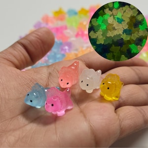 Könnte beinhalten: Eine Hand, die mehrere kleine, farbenfrohe, durchscheinende Tierfiguren hält. Die Figuren sind in den Farbtönen Blau, Rosa, Orange, Weiß und Gelb gehalten. Ein kreisförmiger Einsatz zeigt die grün leuchtenden Figuren. Die Figuren haben schwarze Punktaugen.