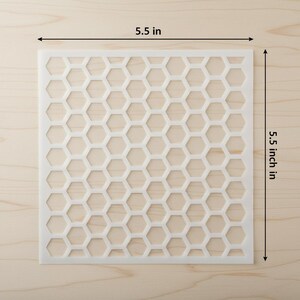 May include: A square, off-white stencil with a honeycomb pattern. The stencil measures 5.5 inches (14 cm) on each side. The design features a repeating pattern of hexagonal shapes, perfect for crafts and DIY projects.