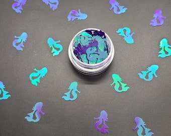 May include: A small clear container filled with iridescent blue and purple mermaid shaped glitter. The glitter is scattered around the container on a black surface.