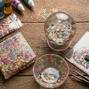 May include: A clear glass snow globe filled with colorful smiley face confetti, with a clear base. A clear bowl and a rectangular container are filled with the same confetti. Additional confetti is scattered on the wooden surface.