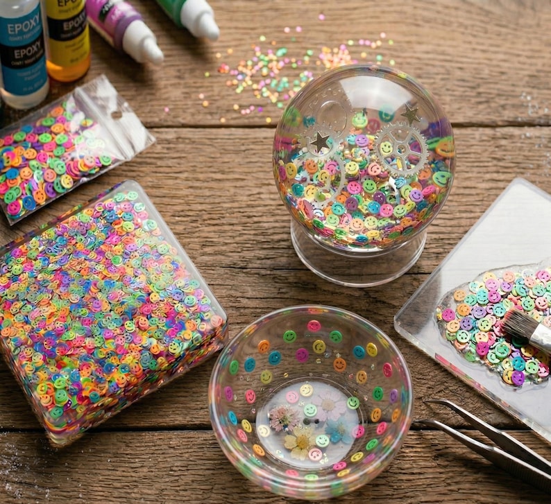 May include: A clear glass snow globe filled with colorful smiley face confetti, with a clear base. A clear bowl and a rectangular container are filled with the same confetti. Additional confetti is scattered on the wooden surface.