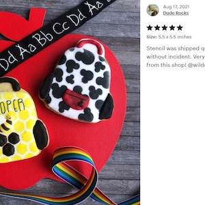 K&ouml;nnte beinhalten: Zwei verzierte Zuckerkekse in Form von Rucks&auml;cken. Ein Keks ist gelb mit schwarzem Wabenmuster und einer Biene, der andere ist wei&szlig; mit schwarzen Flecken und einer roten Tasche. Die Kekse befinden sich auf einer roten, apfelf&ouml;rmigen Oberfl&auml;che mit einem schwarzen Band mit wei&szlig;en Buchstaben, auf dem "a Aa Bb Cc Dd Aa Bb Cc Dd" steht, und einem Regenbogenband.