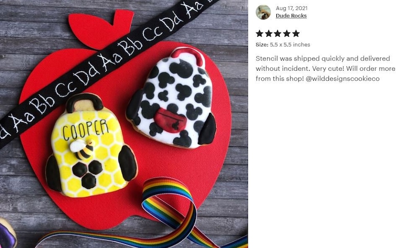 K&ouml;nnte beinhalten: Zwei verzierte Zuckerkekse in Form von Rucks&auml;cken. Ein Keks ist gelb mit schwarzem Wabenmuster und einer Biene, der andere ist wei&szlig; mit schwarzen Flecken und einer roten Tasche. Die Kekse befinden sich auf einer roten, apfelf&ouml;rmigen Oberfl&auml;che mit einem schwarzen Band mit wei&szlig;en Buchstaben, auf dem "a Aa Bb Cc Dd Aa Bb Cc Dd" steht, und einem Regenbogenband.