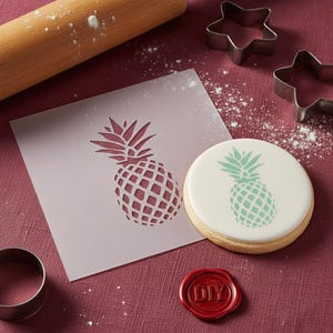 Op de afbeelding: Een bakscène met een ananassjabloon, koekjessnijders en een deegroller. Een glazuurkoekje met een groen ananasontwerp ligt naast de sjabloon. Er is ook een rood zegel met "DIY".