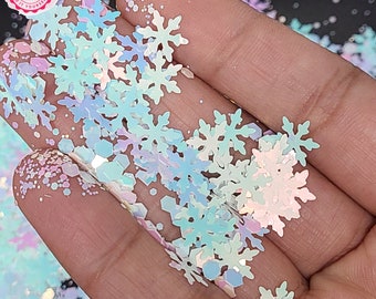 May include: Iridescent snowflake-shaped glitter in various sizes. The glitter has a pearlescent sheen, reflecting shades of blue, green, and pink. The glitter is held in a hand, with some scattered on the surface.