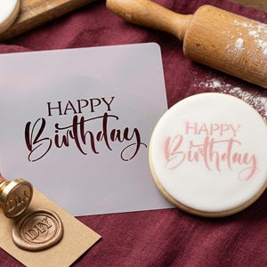 May include: A white frosted cookie with the words "HAPPY Birthday" in pink script. A stencil with the same text is next to the cookie. A rolling pin and other baking tools are in the background.