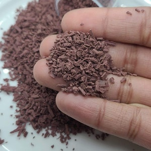 May include: A close-up of a pile of small, brown, shredded pieces of material. The material is in a person's hand, and some of it is scattered on a white surface.