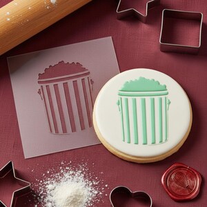 May include: A decorated sugar cookie with a green popcorn bucket design, alongside a stencil, cookie cutters, and a rolling pin. The cookie has a white base with a green popcorn bucket design. A red wax seal is also visible.