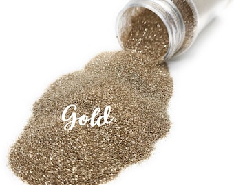 Peut inclure: Un gros plan sur un pot de paillettes dorées, avec le mot "Gold" écrit en blanc sur les paillettes. Les paillettes se répandent du pot sur une surface blanche.