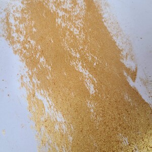 May include: A close-up of a gold glitter paint stroke on a white background. The glitter is dense and creates a textured effect.