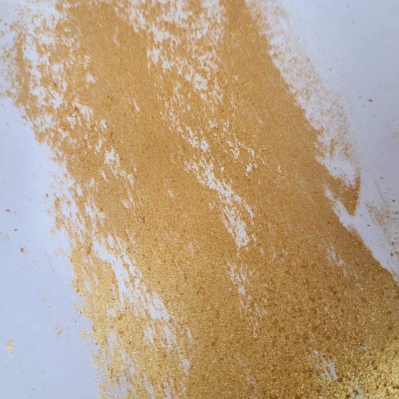 May include: A close-up of a gold glitter paint stroke on a white background. The glitter is dense and creates a textured effect.