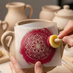 May include: A beige ceramic pot with handles is being decorated with a red and white floral design using a stencil and a sponge brush. The stencil has the words "CUSTOM TEXT" around the edge.