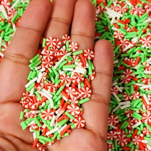 Op de afbeelding: Een close-up van een hand die een verscheidenheid aan rode en groene snoepjesstrooisels vasthoudt. De strooisels zijn in de vorm van snoepstokken en kleine stokjes.