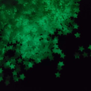 May include: A collection of glowing green star-shaped confetti against a black background. The stars are out of focus, creating a soft, ethereal effect. The image suggests a festive or decorative theme.