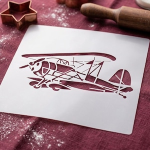 May include: A square, semi-transparent plastic stencil featuring a detailed cutout design of a vintage biplane. The stencil displays the aircraft in profile, showing the wings, propeller, and fuselage structure clearly defined by the negative space.