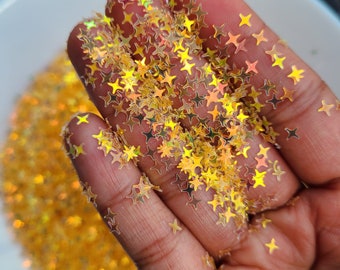 May include: A handful of iridescent gold star-shaped glitter. The glitter is scattered on a white surface.