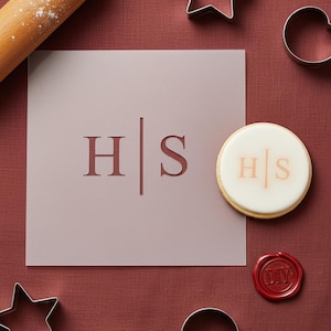 Peut inclure: Un pochoir mauve clair avec les lettres "H" et "S" séparées par une ligne verticale. Un biscuit avec les mêmes initiales est à côté du pochoir. Des emporte-pièces et un rouleau à pâtisserie sont également dans l'image.