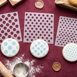 May include: A flat lay of baking equipment on a burgundy surface. Features three polka dot stencils, three cookies with blue polka dots, a rolling pin, flour, and a metal measuring cup. Two wax seals with "DIY" text are also visible.