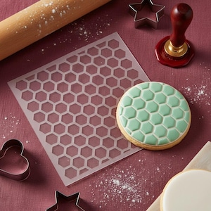 May include: A honeycomb-patterned cookie stencil and a decorated cookie with a green honeycomb design are displayed. Other baking tools include a rolling pin, cookie cutters, and a stamp. The items are arranged on a maroon surface with flour.