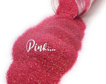 Peut inclure: Gros plan sur une paillettes rose, avec une petite quantité de paillettes dorées mélangées, qui se déversent d'un pot en plastique transparent. Le texte "Pink..." est écrit en blanc sur les paillettes.