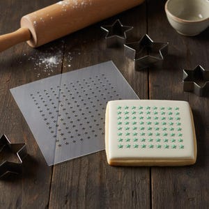 May include: A square cookie decorated with green star sprinkles sits next to a clear stencil with star cutouts. Several star-shaped cookie cutters, a rolling pin, and a small bowl are on a wooden surface.