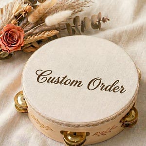 May include: A round tambourine with a cream-coloured head and a light wood frame. The text "Custom Order" is printed on the head in a brown script font. The tambourine has gold-coloured jingles and is decorated with a floral design.