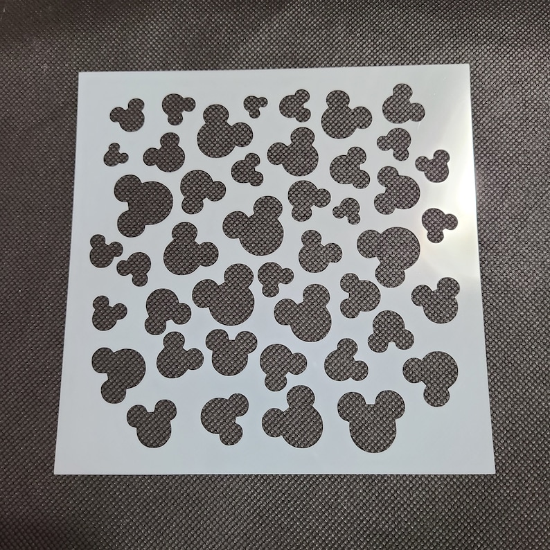 K&ouml;nnte beinhalten: Wei&szlig;e Kunststoffschablone mit einem sich wiederholenden Muster aus Mickey-Maus-K&ouml;pfen in verschiedenen Gr&ouml;&szlig;en.