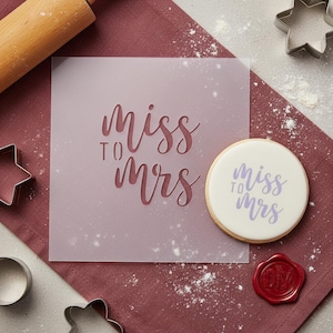 May include: A square stencil with the words "Miss to Mrs" in a script font is next to a decorated sugar cookie with the same text. A rolling pin, cookie cutters, and a red wax seal are also visible.