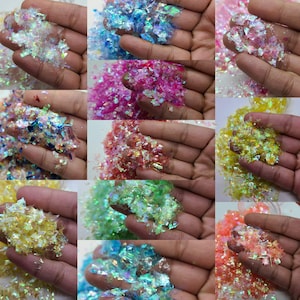May include: Assortment of iridescent glitter flakes in various colors, including white, blue, pink, green, yellow, and orange. The glitter is displayed in small piles on the palms of hands.