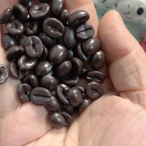 Könnte beinhalten: Nahaufnahme einer Hand, die eine große Menge dunkelbrauner Schokoladenkaffeebohnen hält.