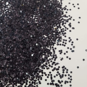May include: A pile of black star-shaped glitter. The glitter is scattered on a white surface.